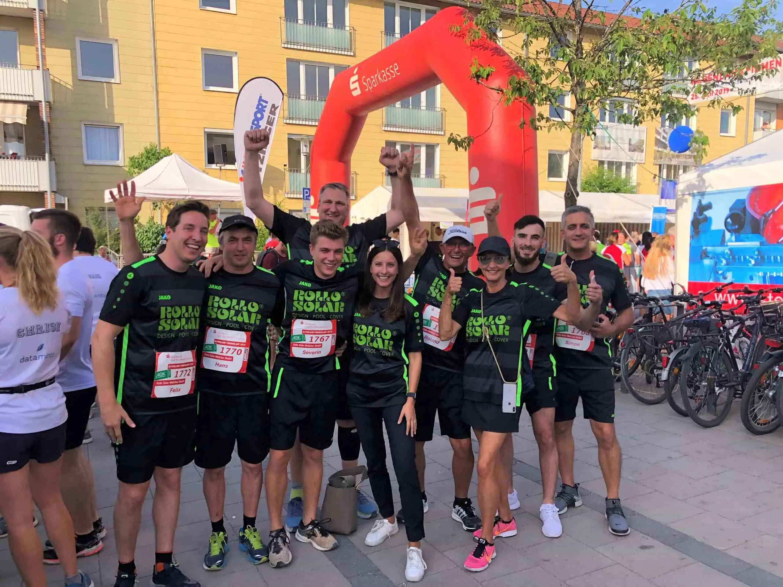 Robustes Team im schwarzen Sportoutfit mit grünen Akzenten beim Laufereignis, jubelnd unter einem großen roten Startbogen, umgeben von Fahrrädern und Zuschauern auf einer Stadtstraße, betont den Gemeinschaftsgeist und sportlichen Erfolg.