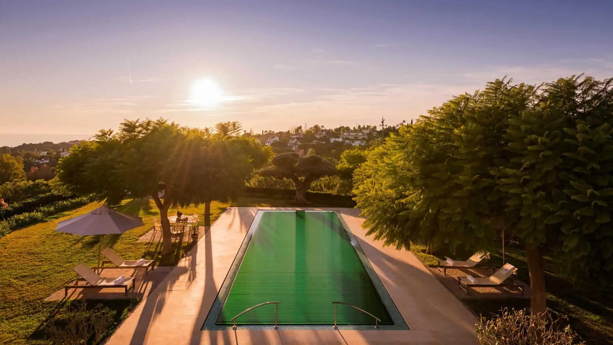 Hochwertiger Poolabdeckung mit Solarenergie im Sonnenlicht, umgeben von Natur und mit komfortablen Liegen und Sonnenschirmen für die perfekte Pool-Atmosphäre.